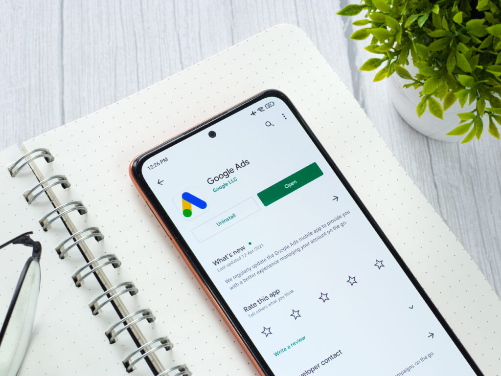 A top-down view of a smartphone resting on a white spiral notebook. The phone screen displays the Google Ads app in the Google Play Store. A pair of glasses and a small green potted plant are visible on a light wood desk in the background.