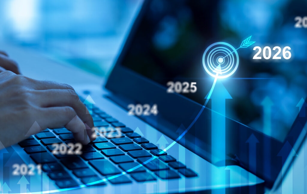 Hands typing on a laptop keyboard with overlaid digital graphics of upward arrows, a timeline from 2021 to 2026, and a glowing target symbol marking 2026 as a goal.