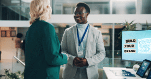 A professional interaction at a conference booth with a digital screen in the foreground displaying "Build Your Brand" and a large, scannable QR code created in Canva.