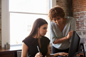 Two female colleagues in a modern office looking intently at a computer screen together, collaborating on a digital marketing strategy for Google and AI search results.