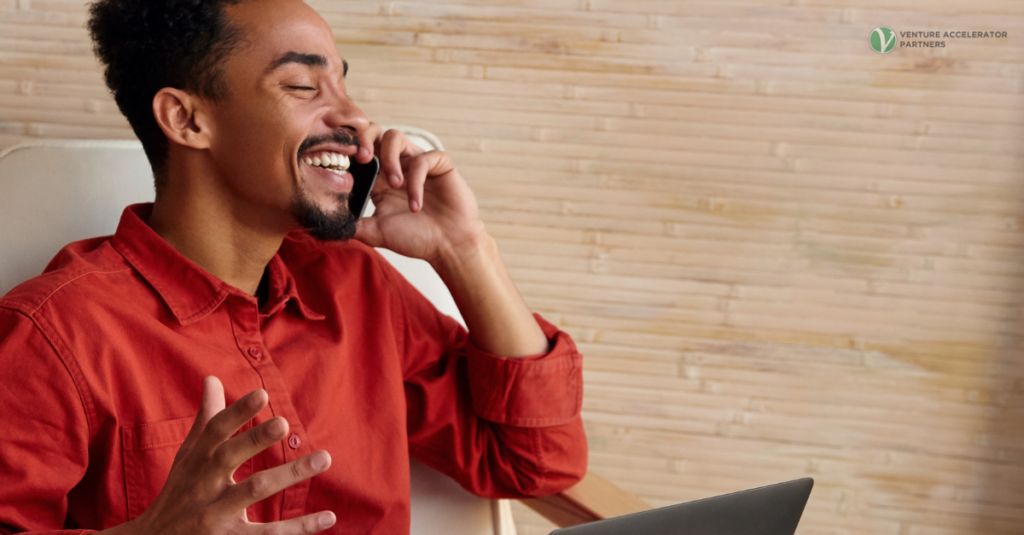 A smiling B2B sales professional wearing a red shirt talking on a phone call with a laptop nearby.