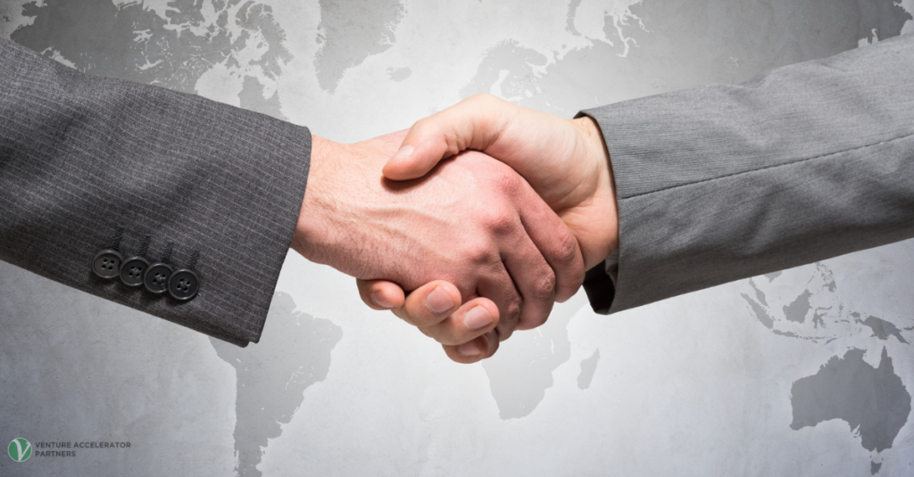 A close-up of two business hands in grey suits shaking hands firmly over a faint grey textured world map. The Venture Accelerator Partners logo is in the lower left.